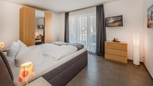 A modern hotel room with a large bed, a wooden wardrobe, a bedside table with a lamp, a dresser, and a painting on the wall.