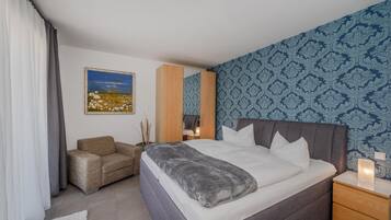 A bedroom with a bed, a chair, a bedside table, a nightstand, and a painting on the wall.