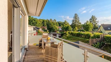 A balcony with a wooden deck, a wicker table with a yellow bowl, and a view of a residential area with houses and trees.