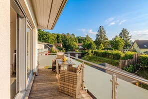 A balcony with a wooden deck, a wicker table with a yellow bowl, and a view of a residential area with houses and trees. - Komfortable, Gemütliche Fewo am Wolgastsee mit Wlan und Kamin. - Villa Petra (Korswandt)