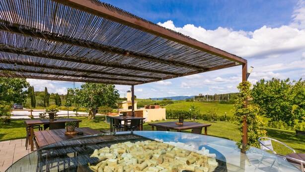 An outdoor patio with a glass-top table, wooden chairs, and a fire pit filled with wine corks. - Podere la Maestà by Interhome (Rosignano Marittimo)