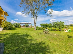 Uma área externa com gramado, uma árvore, uma estrutura coberta e um pequeno jardim.