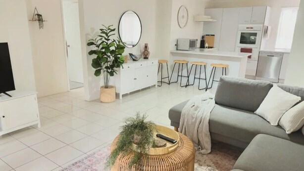 Un salon moderne avec un canapé gris, une table basse ronde en osier agrémentée d’une plante, un téléviseur posé sur un meuble blanc, un coin cuisine avec des meubles blancs et un four à micro-ondes, et un tapis à motifs.