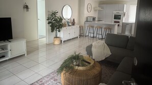 Un salon moderne comprenant un téléviseur, un canapé, une table basse avec une plante, et un coin cuisine équipé d’un four à micro-ondes, d’un four et de tabourets de bar.