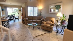 A spacious living room with a ceiling fan, a large window, a sofa, a coffee table, and a dining area with chairs and a table.