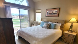 A bedroom with a large bed, a ceiling fan, a nightstand with a lamp, and a window with a view of a golf course.