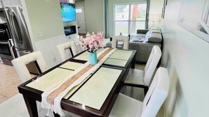 A modern dining area with a long table, white chairs, and hanging pendant lights. - Seabreeze garden oasis, jacuzzi, golf, mins ->beaches (Largo)