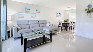 A modern living room with a grey sofa, a glass coffee table, and a dining area with a white table and chairs. - Seabreeze garden oasis, jacuzzi, golf, mins ->beaches (Largo)