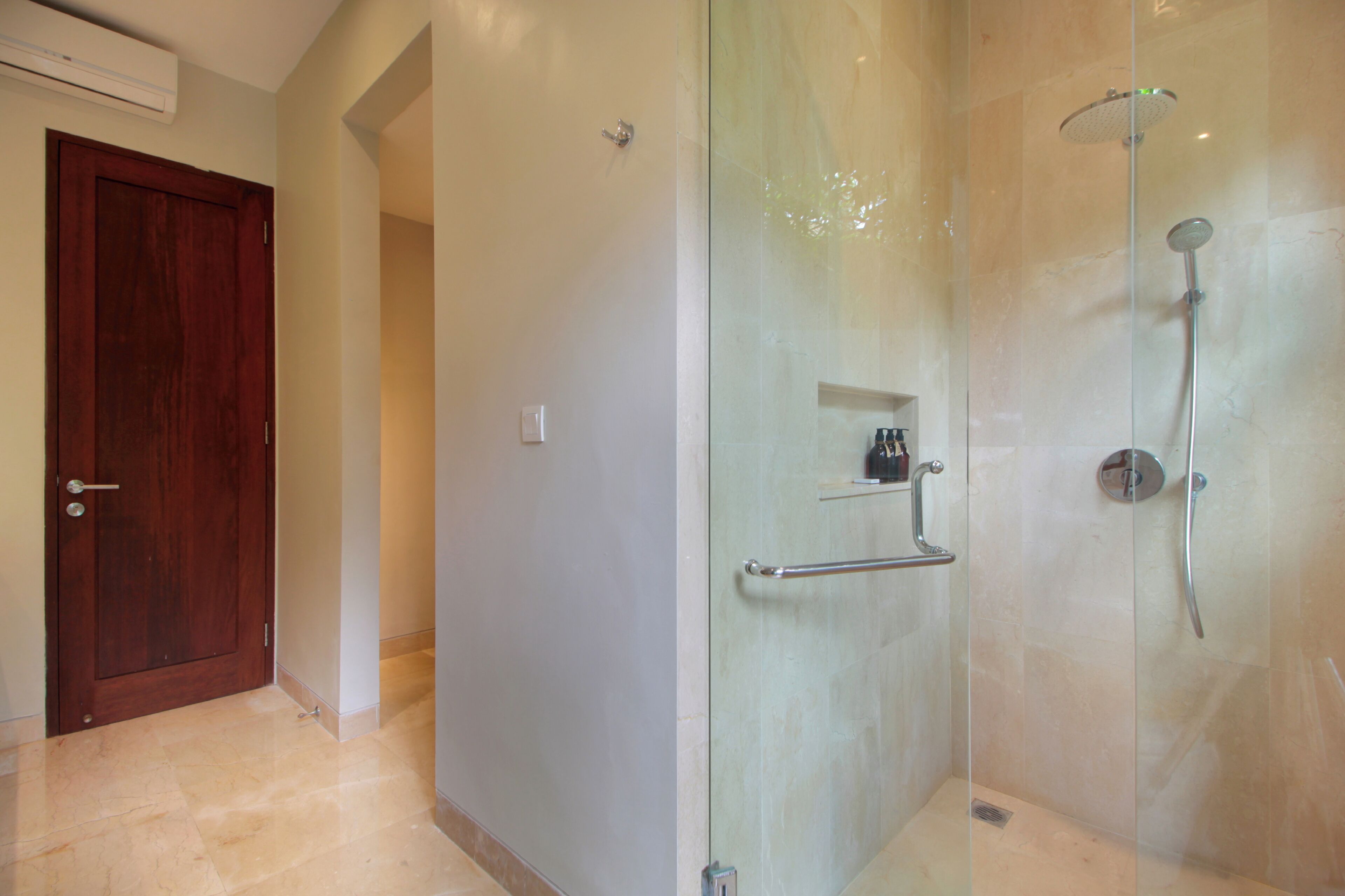 A bathroom with a glass shower enclosure, a wooden door, and a small shelf with toiletries.