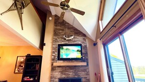 A living room with a fireplace, a ceiling fan, a television, and a large window. - Not Your Everyday Mini Chalet, foot of Holiday Valley Resort! Walk to Downtown (Ellicottville)