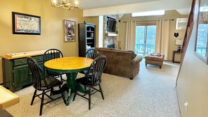 A dining area with a round table and chairs, a sofa, and a television mounted on a stone wall. - Not Your Everyday Mini Chalet, foot of Holiday Valley Resort! Walk to Downtown (Ellicottville)