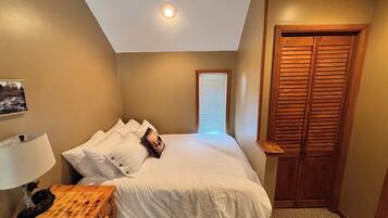 Ein Schlafzimmer mit einem Bett, einem Nachttisch, einer Lampe, einer hölzernen Bank, einem Fenster mit Jalousien und einer Holztür.