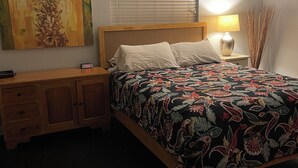 Un dormitorio con cama, mesita de noche, lámpara, ventilador de techo, un cuadro en la pared y una ventana con persianas.