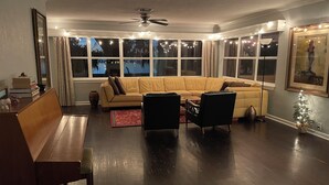 Una sala de estar con un amplio sofá en forma de L, una mesa de centro y dos sillones. Hay varias ventanas con cortinas, un ventilador de techo y un pequeño árbol de Navidad.