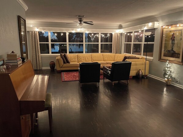 Un salon comprenant un grand canapé en forme de L, une table basse et deux fauteuils. On y trouve plusieurs fenêtres avec des rideaux, un ventilateur de plafond et un petit sapin de Noël.