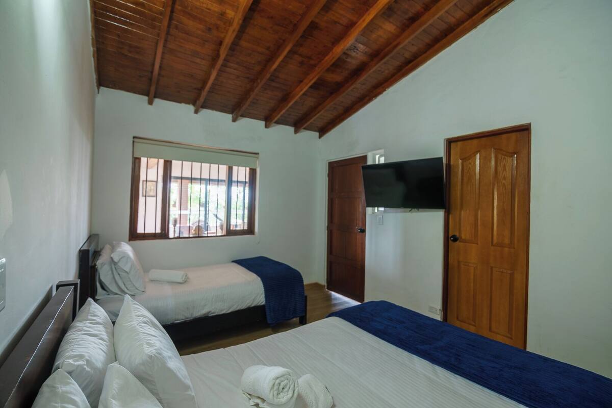 Une chambre avec deux lits, un plafond en bois, une fenêtre avec des barreaux, un téléviseur fixé au mur et une porte en bois.