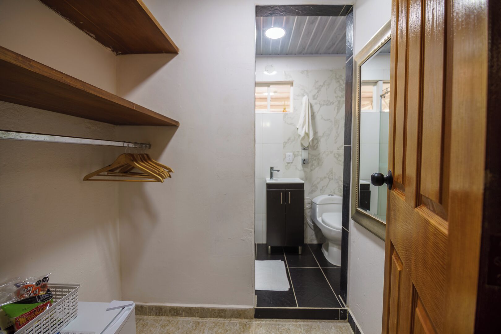 Un couloir étroit mène à une salle de bain équipée de toilettes, d’un lavabo et d’un miroir. On y trouve des étagères en bois et un panier, situés sur la gauche.