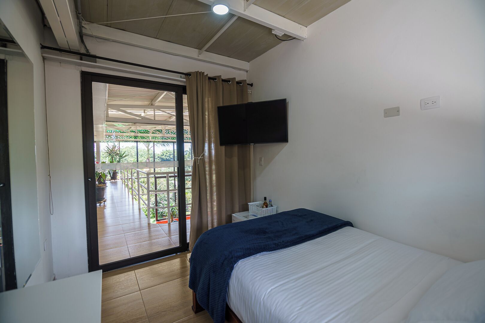 Habitación de hotel con cama, televisión montada en la pared, balcón con vista a zonas verdes y puerta que da a un espacio al aire libre.