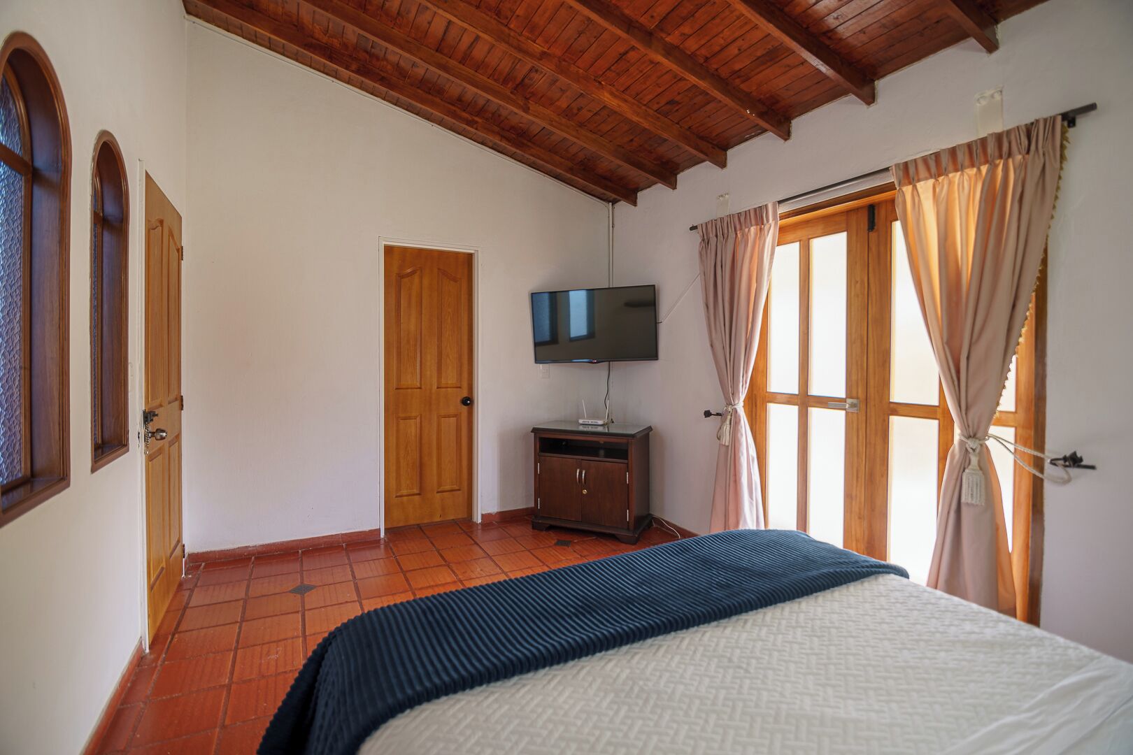 Un dormitorio con techo de madera, una cama, un televisor sobre un mueble y una ventana con cortinas.