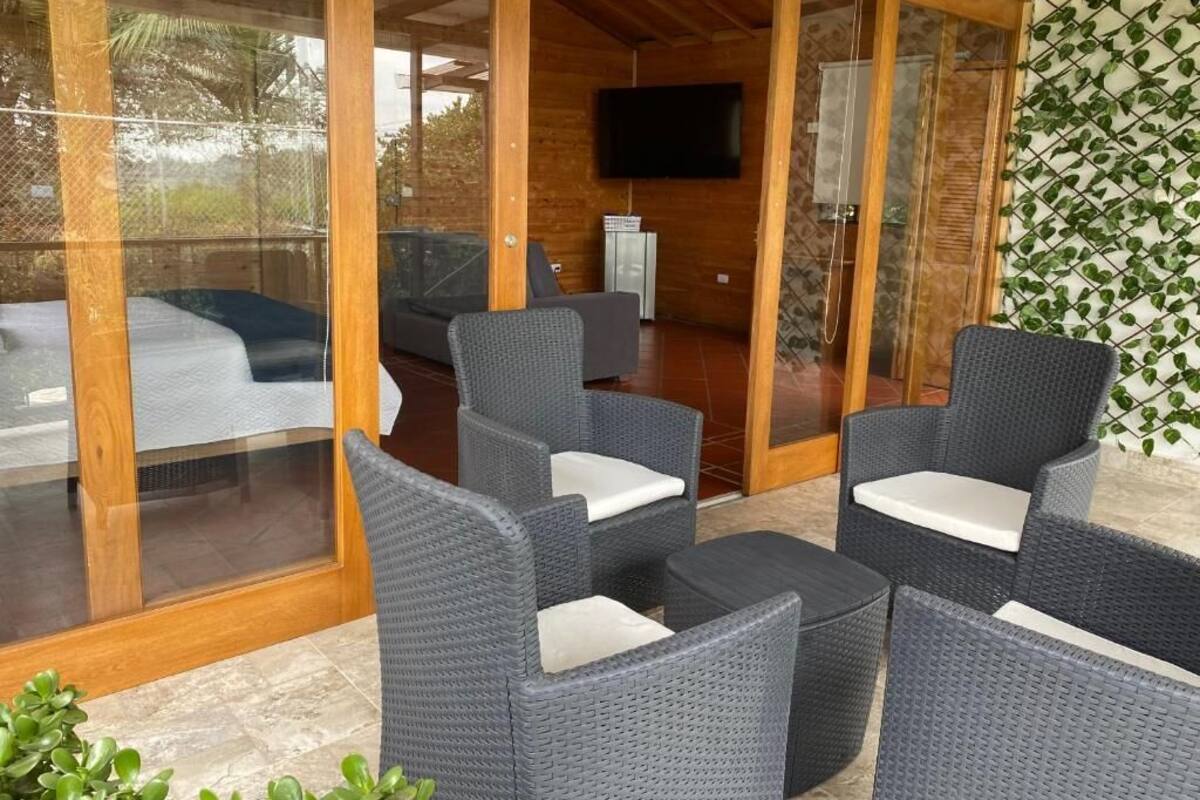 Un espace extérieur moderne, aménagé avec des fauteuils en osier et une petite table, un téléviseur fixé à un mur en bois, et une porte vitrée donnant sur un patio couvert.