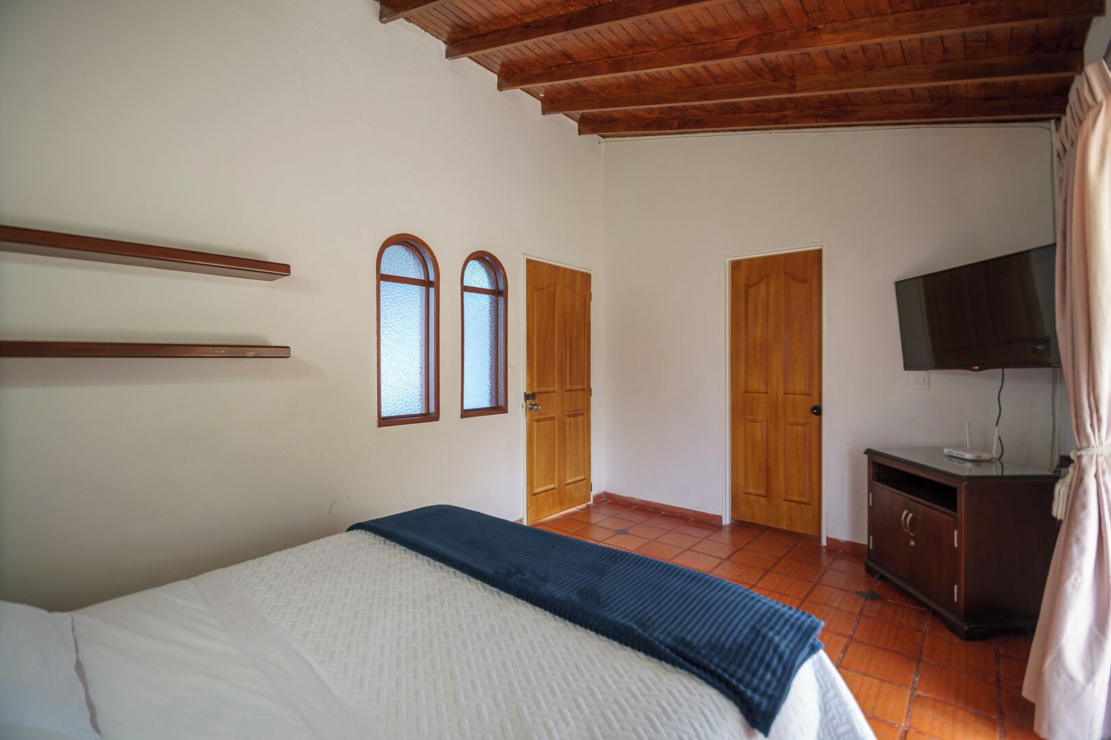 A bedroom with a bed, wooden doors, a television, and a tiled floor.