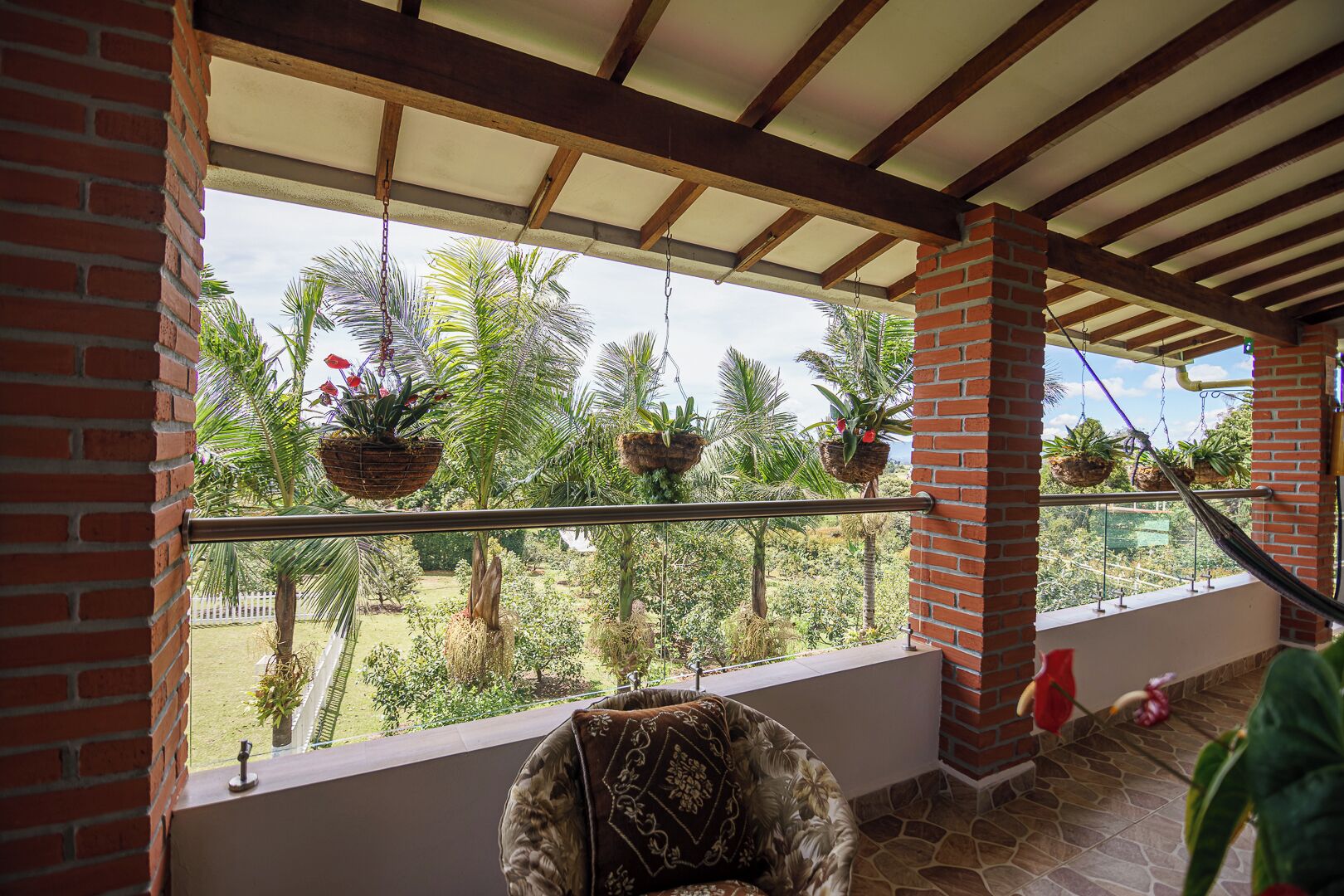 A covered balcony with hanging planters, a cushioned chair, and a view of a lush garden.