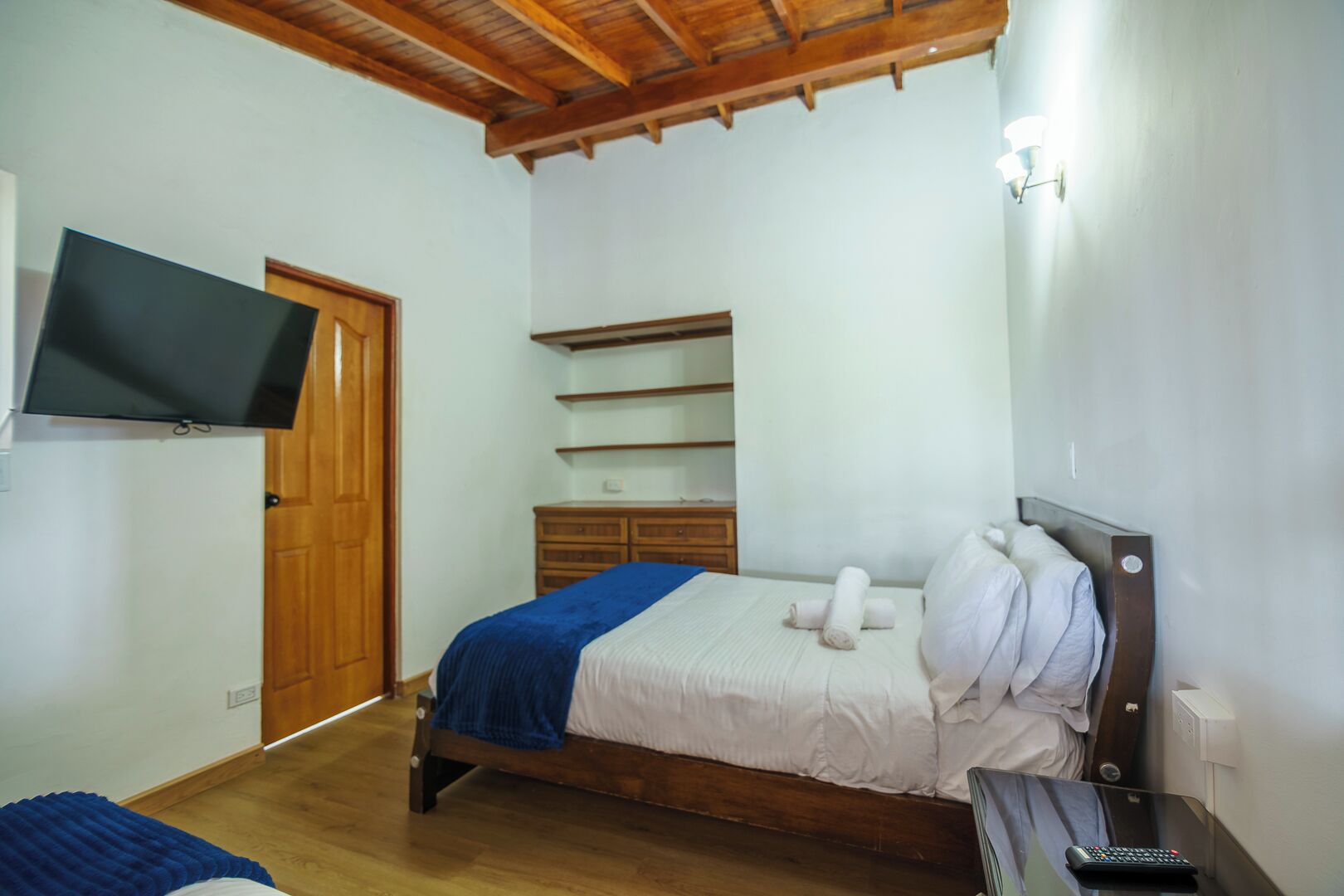 Une chambre avec un lit en bois, un téléviseur fixé au mur, une commode en bois et une étag ère.