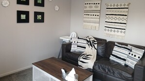 Una sala de estar con un sofá de cuero marrón, una silla blanca y una mesa de centro de madera. Hay cuatro cuadros enmarcados en la pared y dos mantas estampadas.