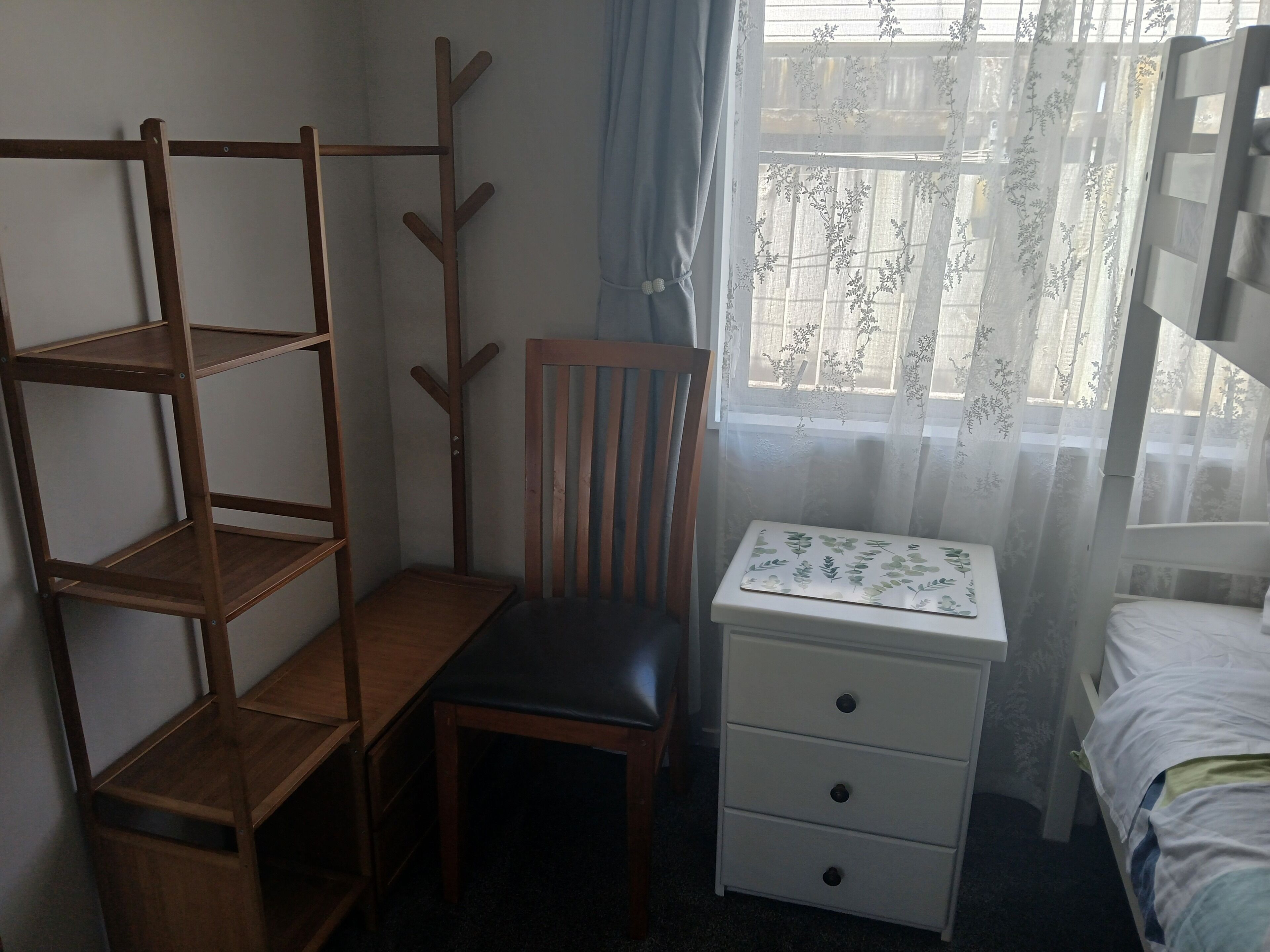 Makuuhuoneessa on puinen tuoli, yöpöytä laatikoilla, pieni hyllykkö ja ikkuna, jossa on ohuet verhot.