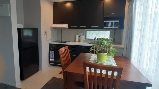 Cocina compacta con armarios oscuros, un refrigerador negro y una mesa de comedor con sillas. Hay una planta en maceta sobre la mesa, y se ve una ventana con cortinas.