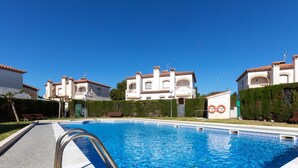 En swimmingpool med en klar, blå vandoverflade, omgivet af en hvid poolterrasse. I baggrunden ses hvide bygninger med røde tegltage og flere altaner.