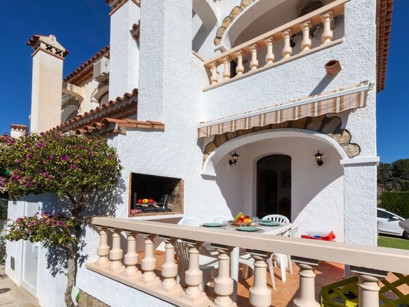 A white building with a balcony, a stone chimney, and a window with a table set for dining.