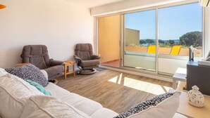 Una sala de estar moderna con un sofá, dos sillones y una mesita. Unos ventanales corredizos dan a un espacio exterior con sillas amarillas.