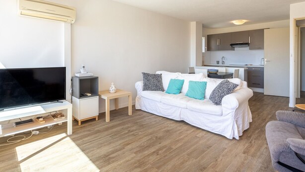 Una sala de estar moderna con un televisor de pantalla plana, un sofá blanco con cojines estampados, una mesita auxiliar y un sillón gris.
