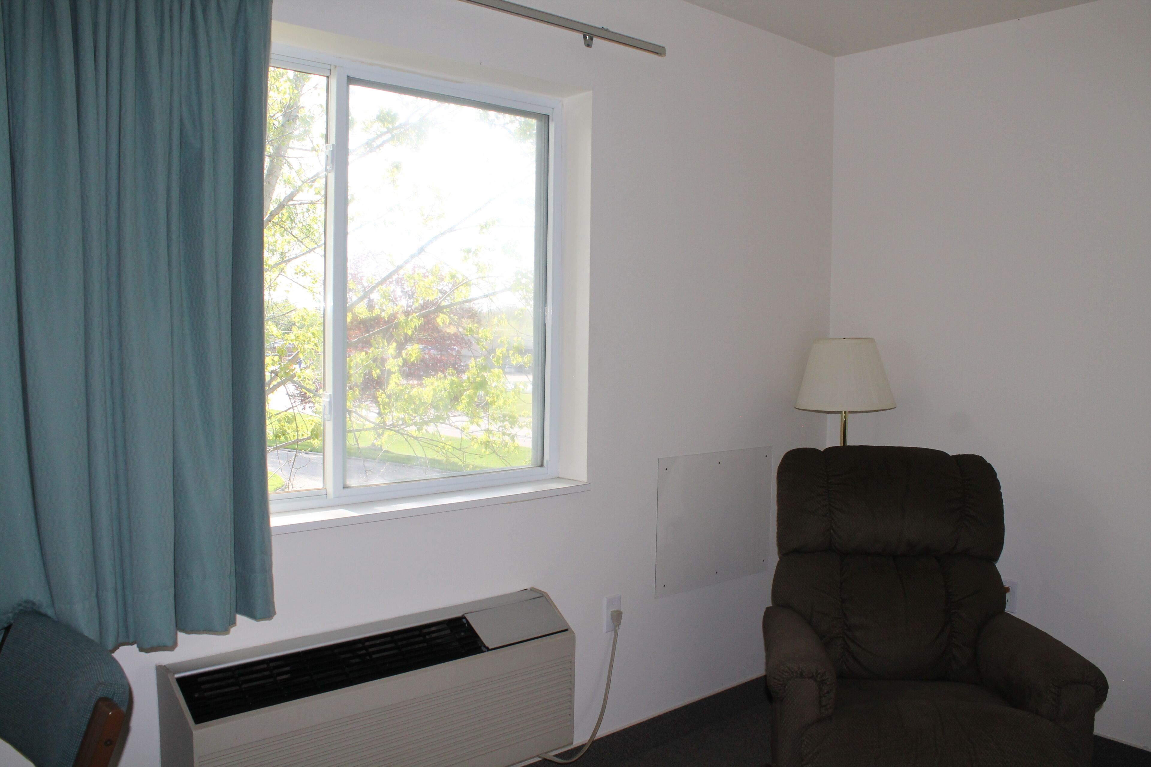 A room with a window, an air conditioning unit, a chair, and a lamp.