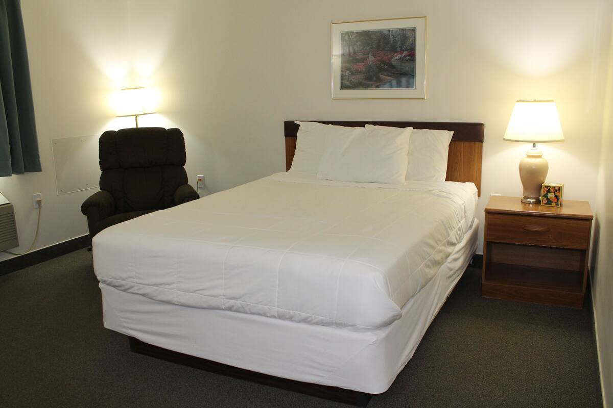 Una habitación de hotel con una cama grande, una silla, una mesita de noche con lámpara y un cuadro en la pared.