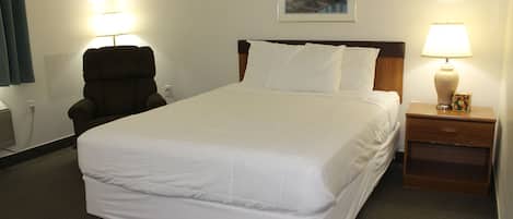 Una habitación de hotel con una cama grande, una silla, una mesita de noche con lámpara y un cuadro en la pared.