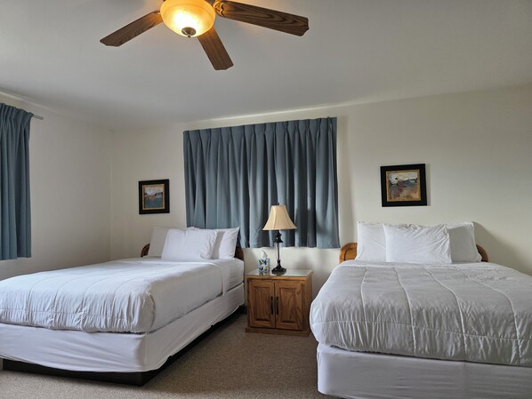 A hotel room with two beds, a ceiling fan, a nightstand with a lamp, and two framed pictures on the walls. - American Host Inn (Hart)