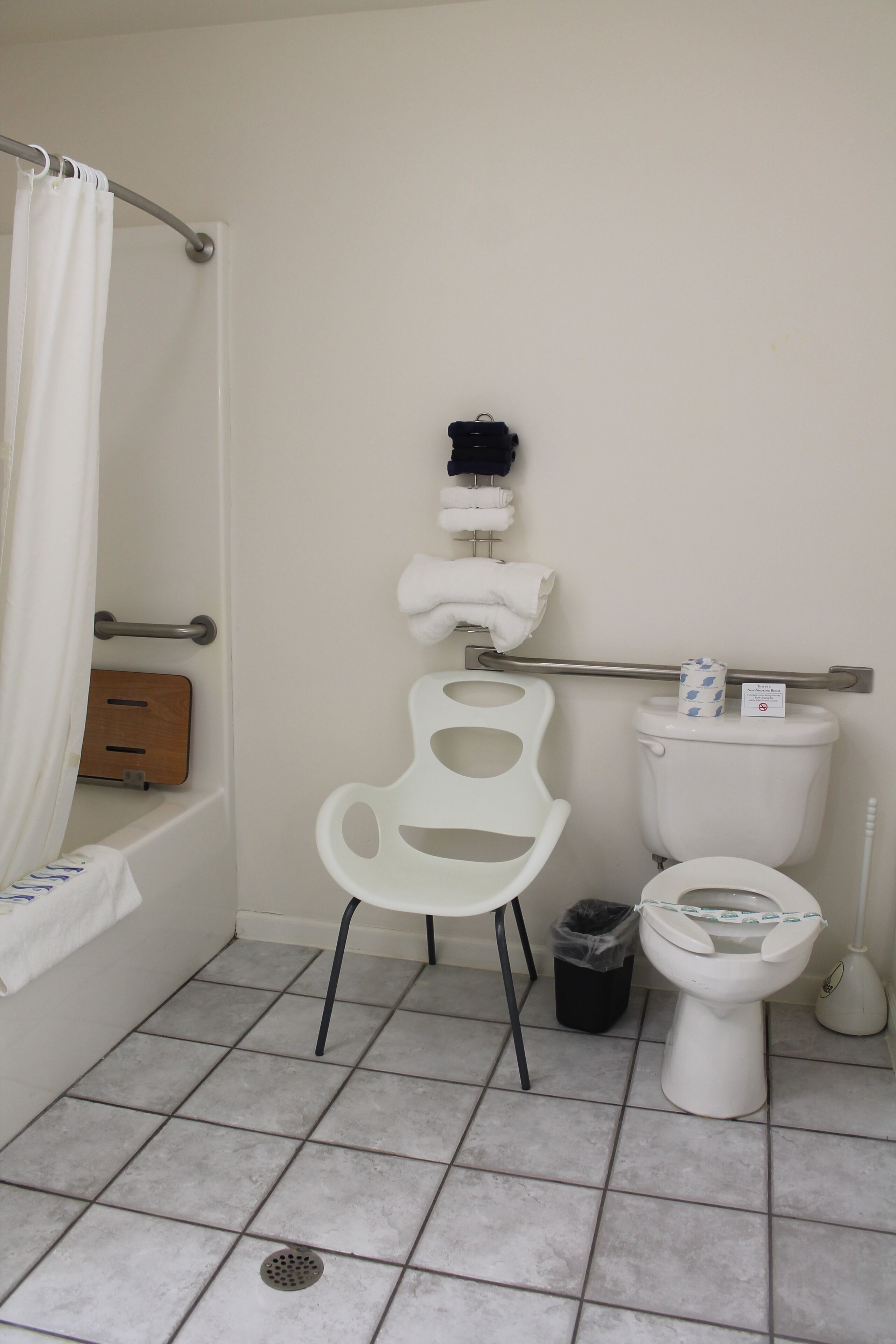 A bathroom with a white toilet, a white sink, a shower with a white curtain, a grab bar, and a trash bin.