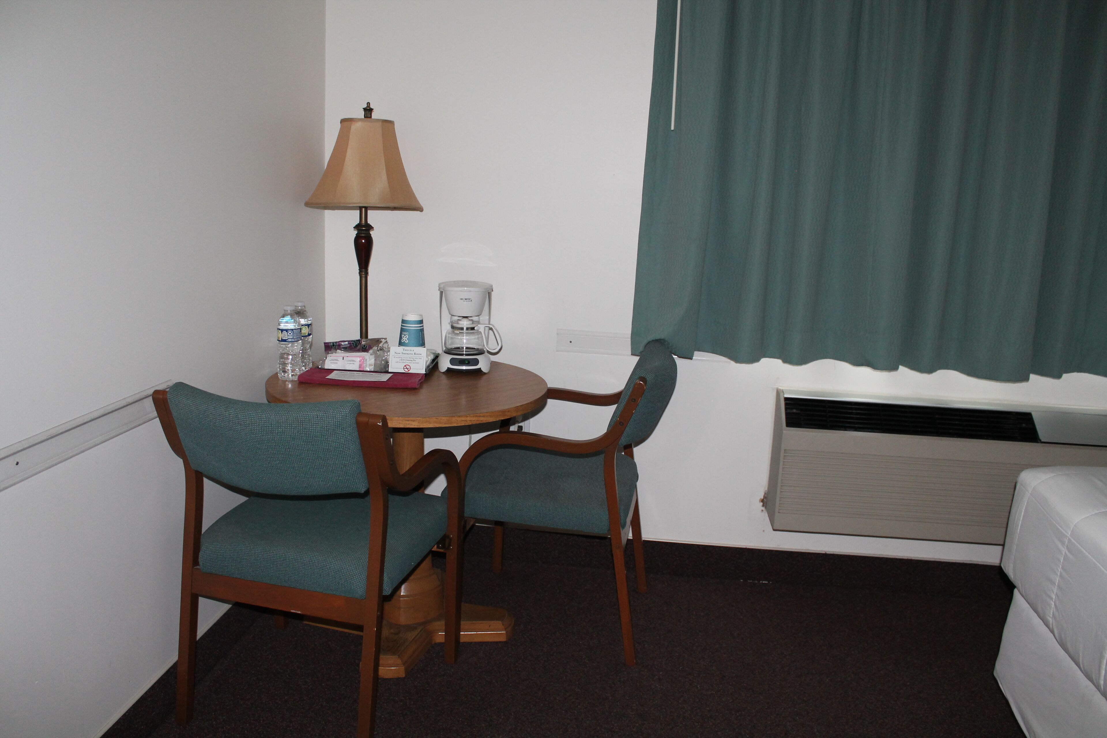 Ein Hotelzimmer mit einem kleinen runden Tisch, zwei Stühlen, einer Lampe, einer Kaffeemaschine und einem Fenster mit Vorhängen.