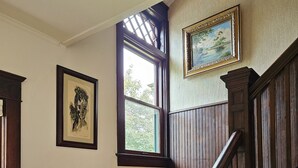 Een houten trap met een leuning, een raam met uitzicht op bomen, en twee ingelijste kunstwerken aan de muur.