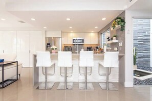 A modern kitchen with white cabinets, a stainless steel refrigerator, a microwave, and a coffee machine. There are four white bar stools in front of the kitchen.