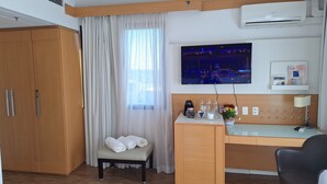 A hotel room with a wooden wardrobe, a bench with towels, a desk with a coffee maker, glasses, and a flower vase. - HOTEL FARIA LIMA (São Paulo)
