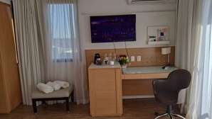 A hotel room with a wooden desk, a chair, a bench with towels, a television on the wall, and a window with curtains. - HOTEL FARIA LIMA (São Paulo)