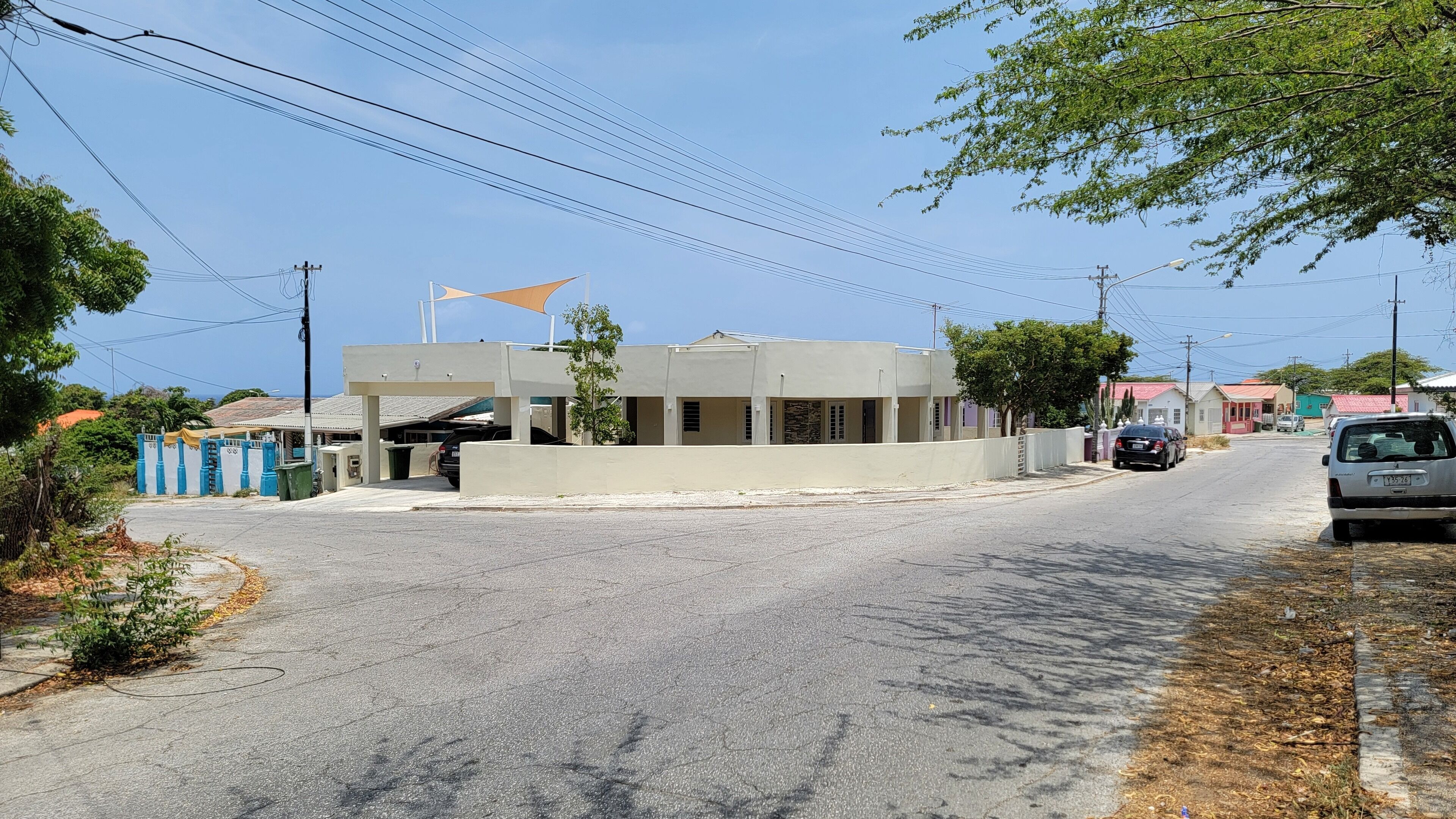 A street view with a building that has a flat roof and a single-story structure. There are cars parked along the street, and a few trees are visible.