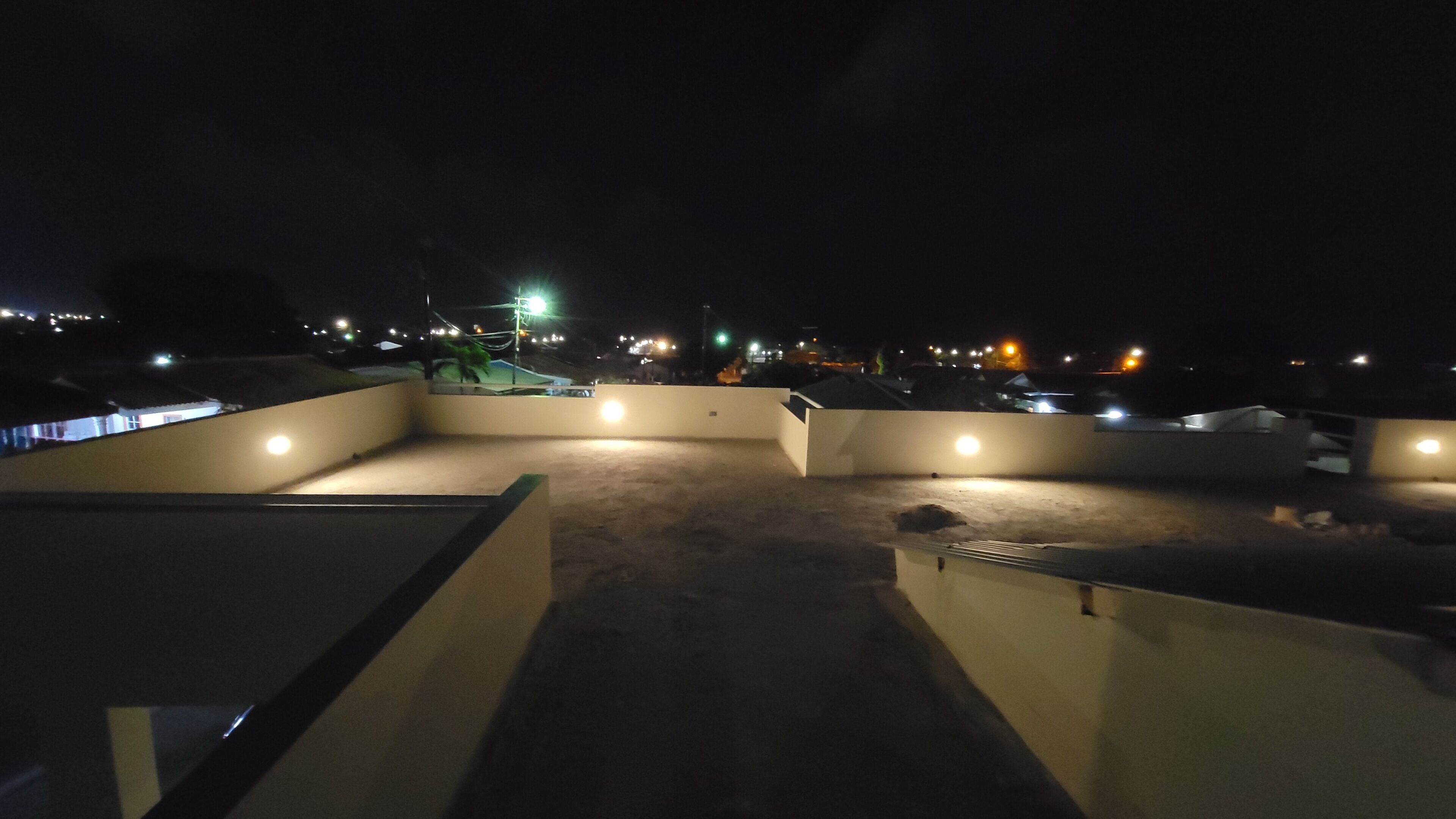 A rooftop terrace with multiple light fixtures, a concrete floor, and a view of the surrounding area at night.