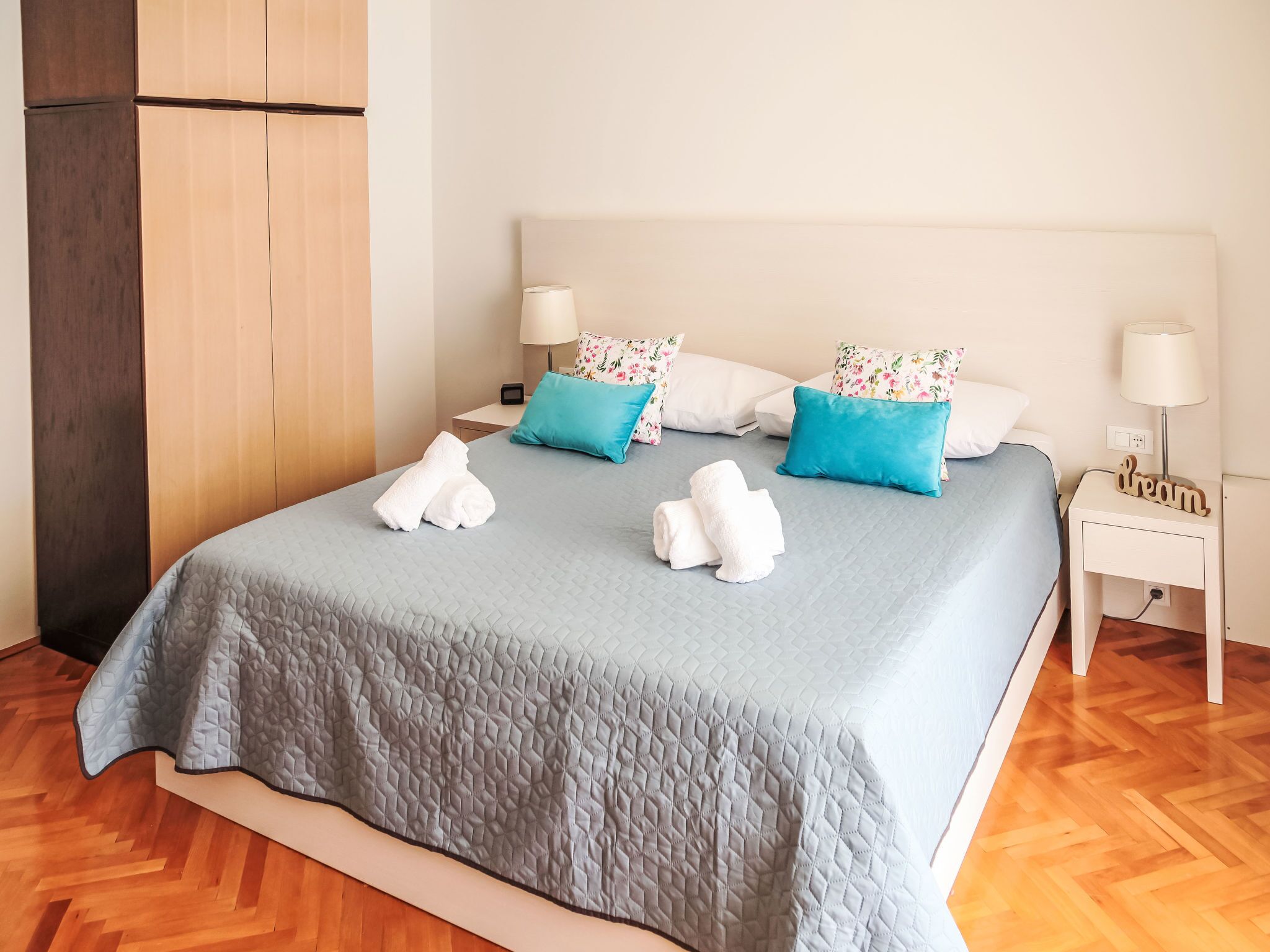 Ein ordentlich gemachtes Bett mit einer gesteppten, grauen Decke, zwei weißen Handtüchern und blumigen Kissen. Es gibt zwei Nachttische mit Lampen und einen kleinen Holz-Nachttisch mit einem 'dream'-Schild.