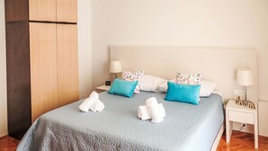 Ein ordentlich gemachtes Bett mit einer gesteppten, grauen Decke, zwei weißen Handtüchern und blumigen Kissen. Es gibt zwei Nachttische mit Lampen und einen kleinen Holz-Nachttisch mit einem 'dream'-Schild.