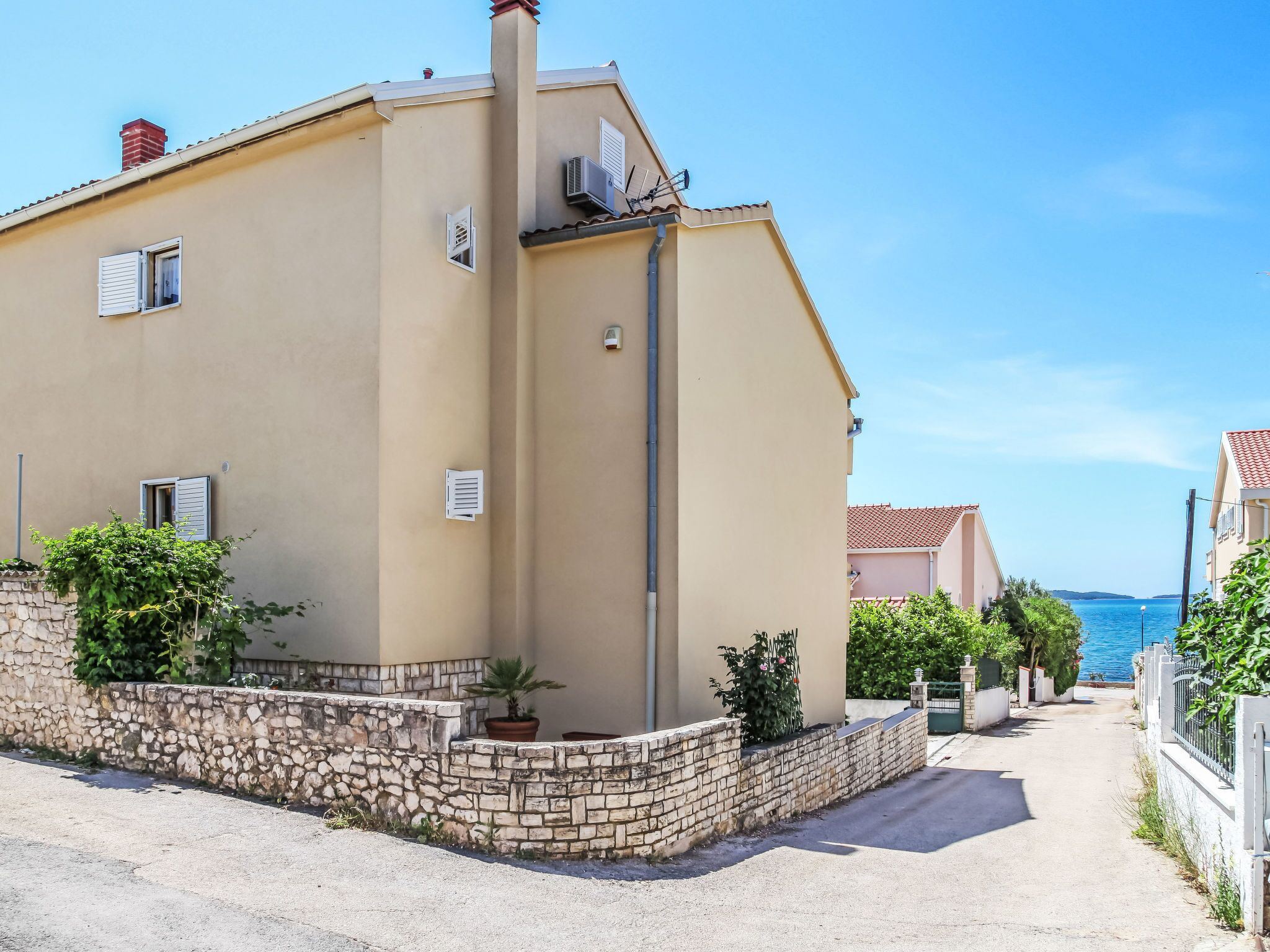 A two-story building with a beige facade, a stone wall at the base, and a clear view of the sea in the background.