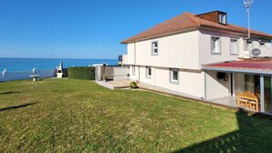 Une maison à deux étages, avec un toit recouvert de tuiles rouges, un balcon agrémenté d’une table et de chaises, et une vue sur la mer.